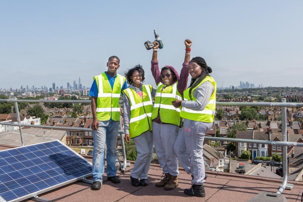 Four people stand on a rooftop in London next to some solar panels. They are wearing hi-vis jackets and smiling in the sunshine. Image credit: Repowering London.