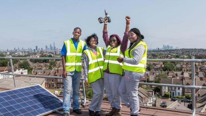 Image credit: Repowering London. Image description: Four people stand on a rooftop in London next to solar panels. They are smiling in the sunshine.